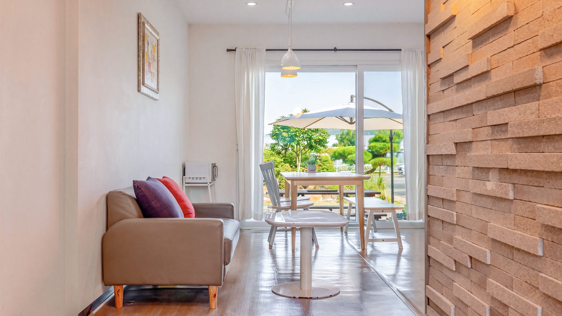 A modern living room with a sofa, a dining table, and chairs, a large window with a view of greenery, and a brick accent wall.