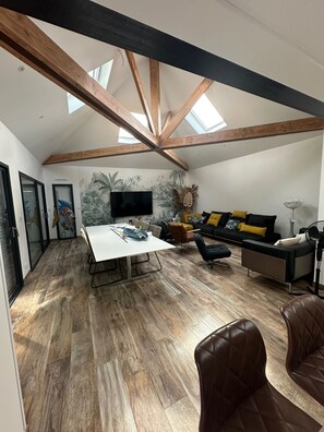 Amplia sala de estar con techo alto, vigas de madera, televisor de pantalla plana, sofá con cojines y una mesa de comedor.