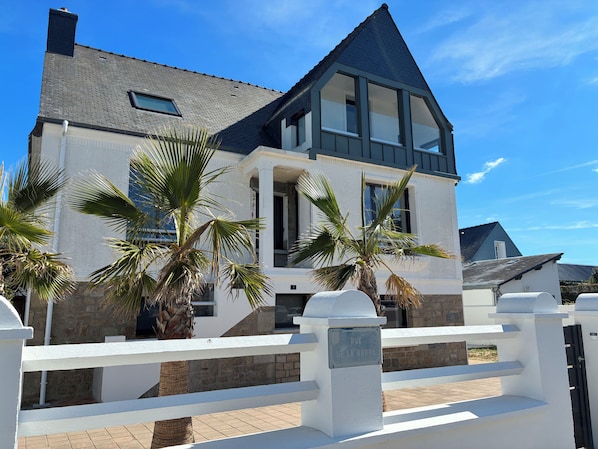 A modern two-story house with a balcony, palm trees, and a white fence.