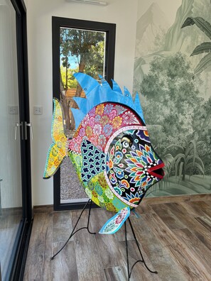 Una escultures de peix colorat sobre un pedestal en una habitació amb una porta i un mural d'un arbre i una muntanya.