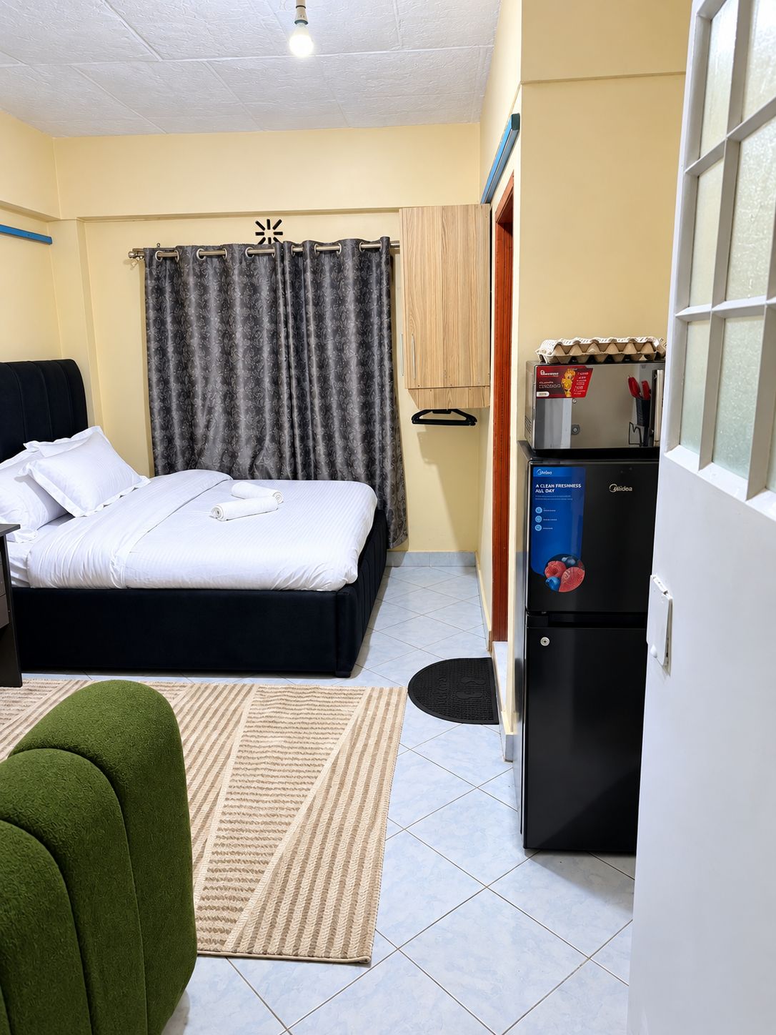 A hotel room with a bed, a small desk, a refrigerator, and a green chair.