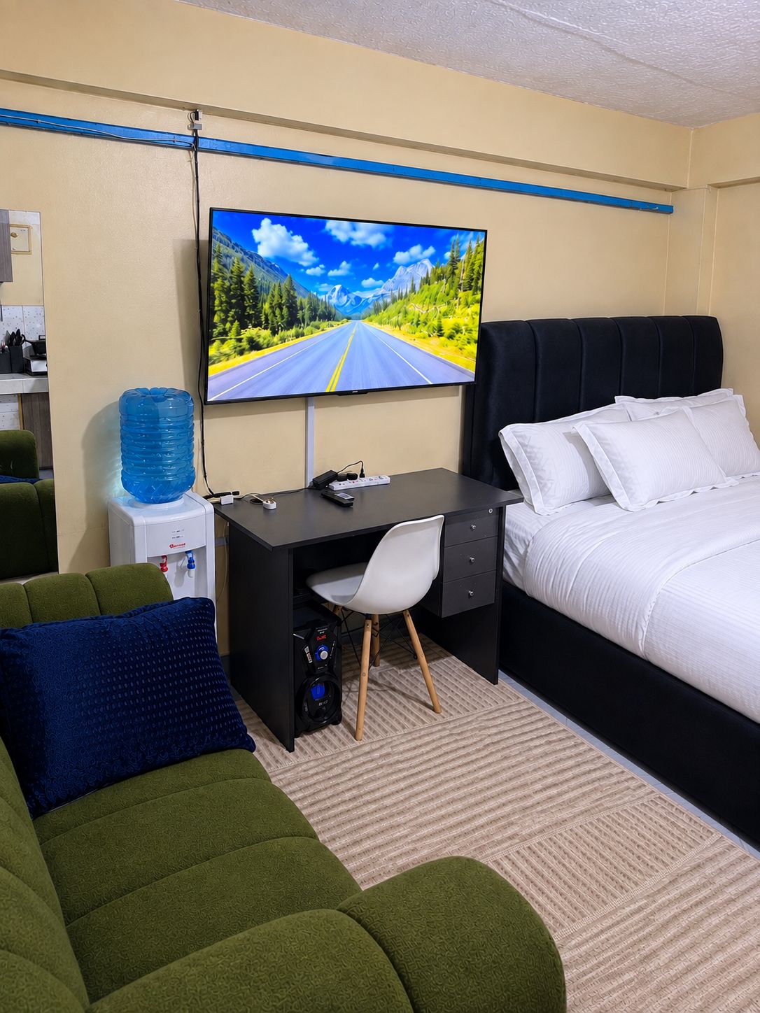 Ein Hotelzimmer mit Bett, Schreibtisch mit Computer, Fernseher zeigt eine malerische Straße und Wasserspender.