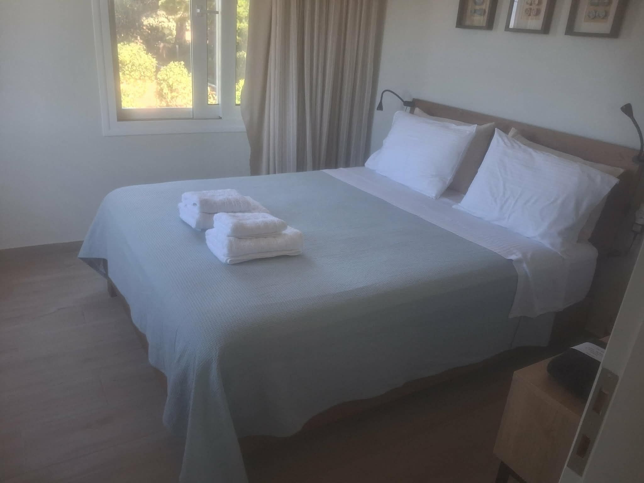 Ein ordentlich gemachtes Bett mit weißen Leinen und einem gefalteten Handtuch, ein Fenster mit Vorhängen und eine Wand mit gerahmten Bildern.