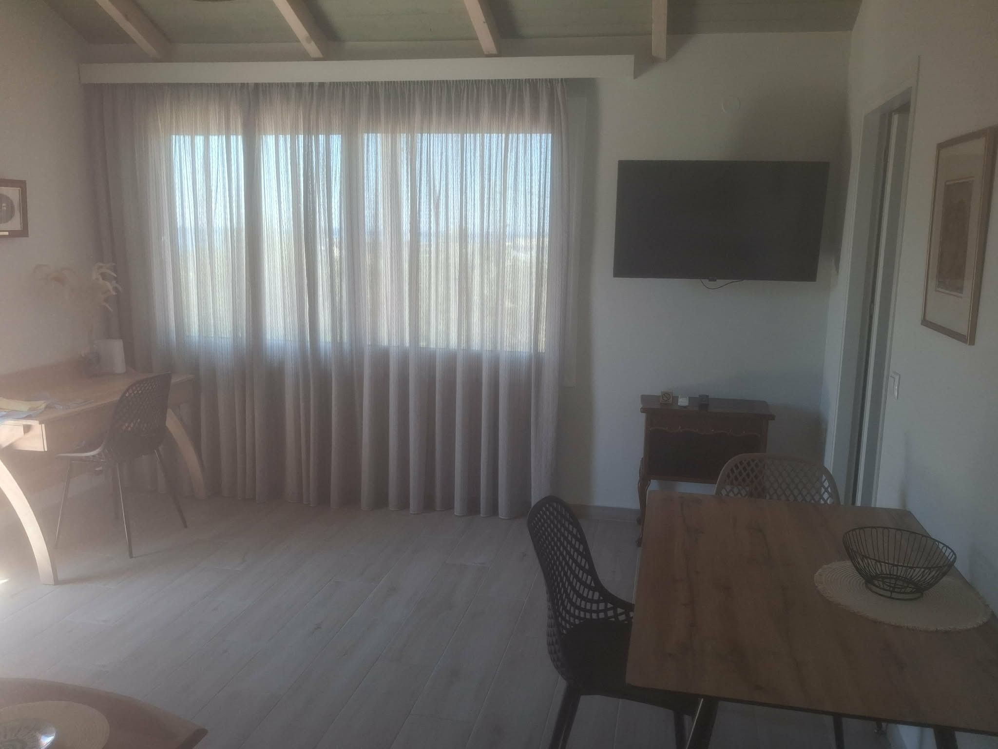 A room with a wooden dining table, chairs, a TV mounted on the wall, and a large window with curtains.