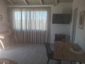 Ein Zimmer mit einem Holztisch, Stühlen, einem an der Wand befestigten Fernseher und einem großen Fenster mit Vorhängen.