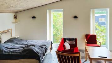 A bedroom with a bed, a red chair, a table, and two windows with a view of greenery.