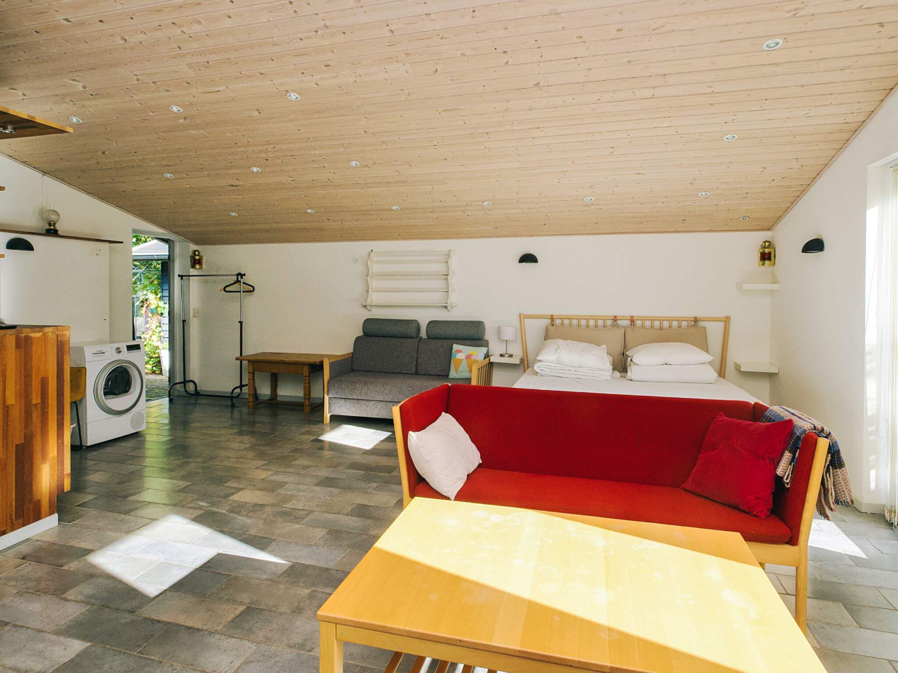 A compact room with a bed, a sofa, a table, a washing machine, and a small table with a lamp.