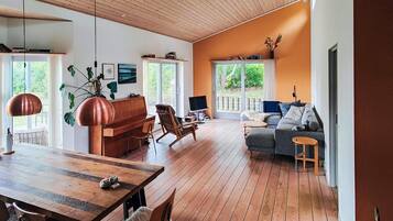 A spacious living room with a wooden floor, a piano, a sofa, and a dining area with a table and chairs.
