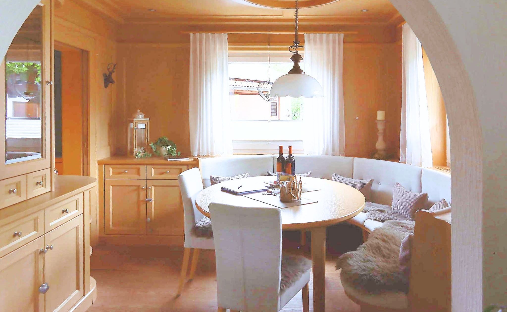 A cozy dining area with a round table, chairs, and a bench. There is a window with curtains, a chandelier, and a cabinet with drawers.