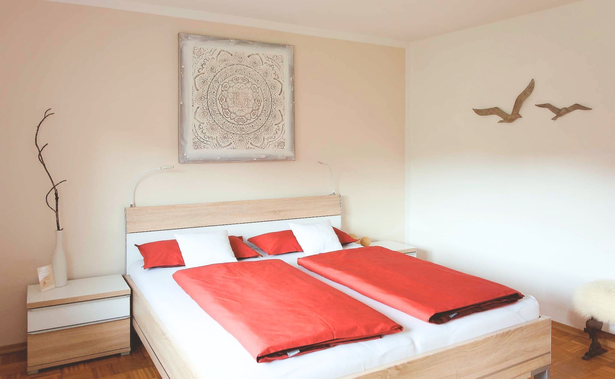 Une chambre à coucher avec un lit en bois, une literie rouge et blanche, une table de chevet, un vase avec une branche, une œuvre d’art murale et une décoration murale représentant des oiseaux.