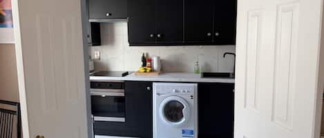 Une cuisine compacte avec des meubles noirs, un four à micro-ondes encastré, un évier et un lave-linge à chargement frontal.