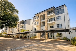 Un edifici d'apartaments de diversos pisos amb balcons, una zona d'entrada pavimentada i mobiliari exterior cobert.