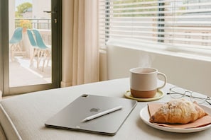 Un ordinador portàtil, una tassa de cafè, una parella de lents i un croissant damunt d'una taula a peu d'una finestra amb persianes.
