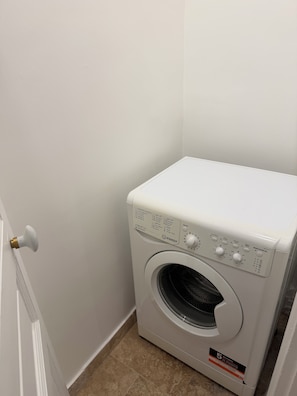 A compact, white front-loading washing machine with a control panel, located in a corner with a white door and tiled floor.