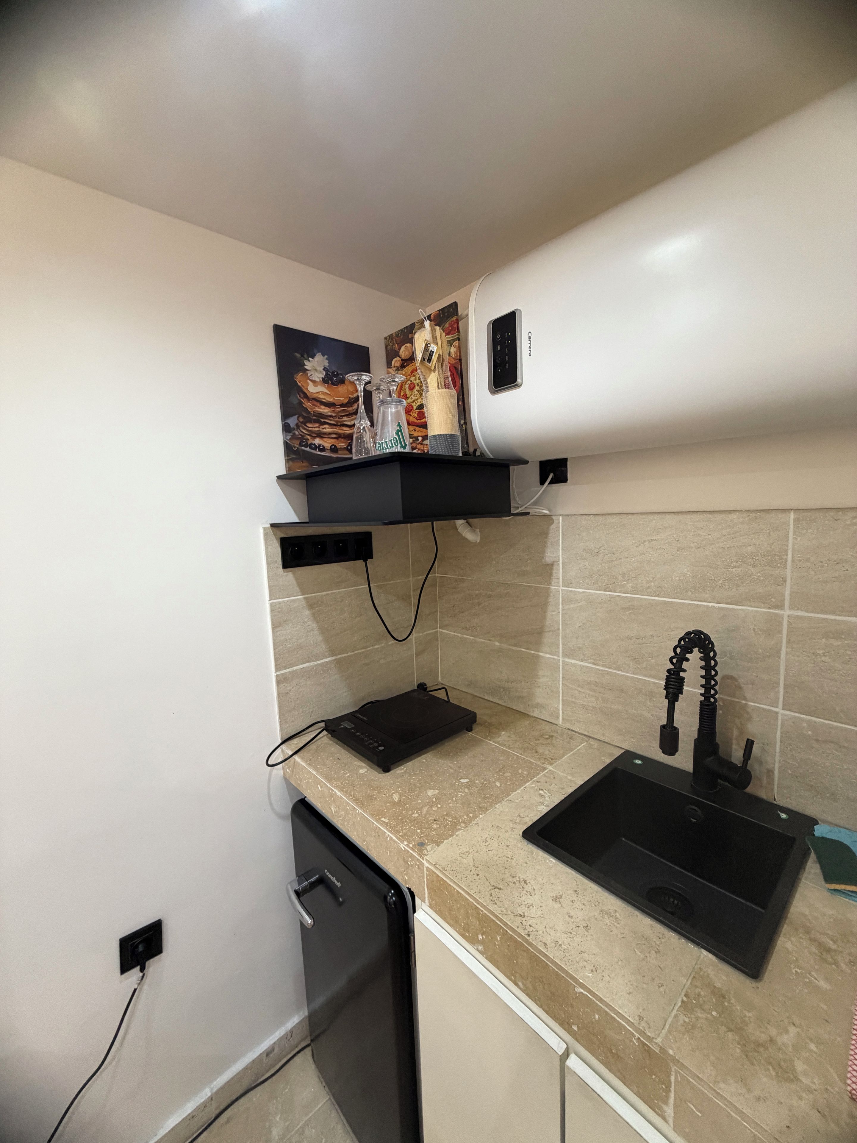 Cocina compacta con encimera negra, un lavabo, lavavajillas y un estante pequeño con un cuadro y una botella.