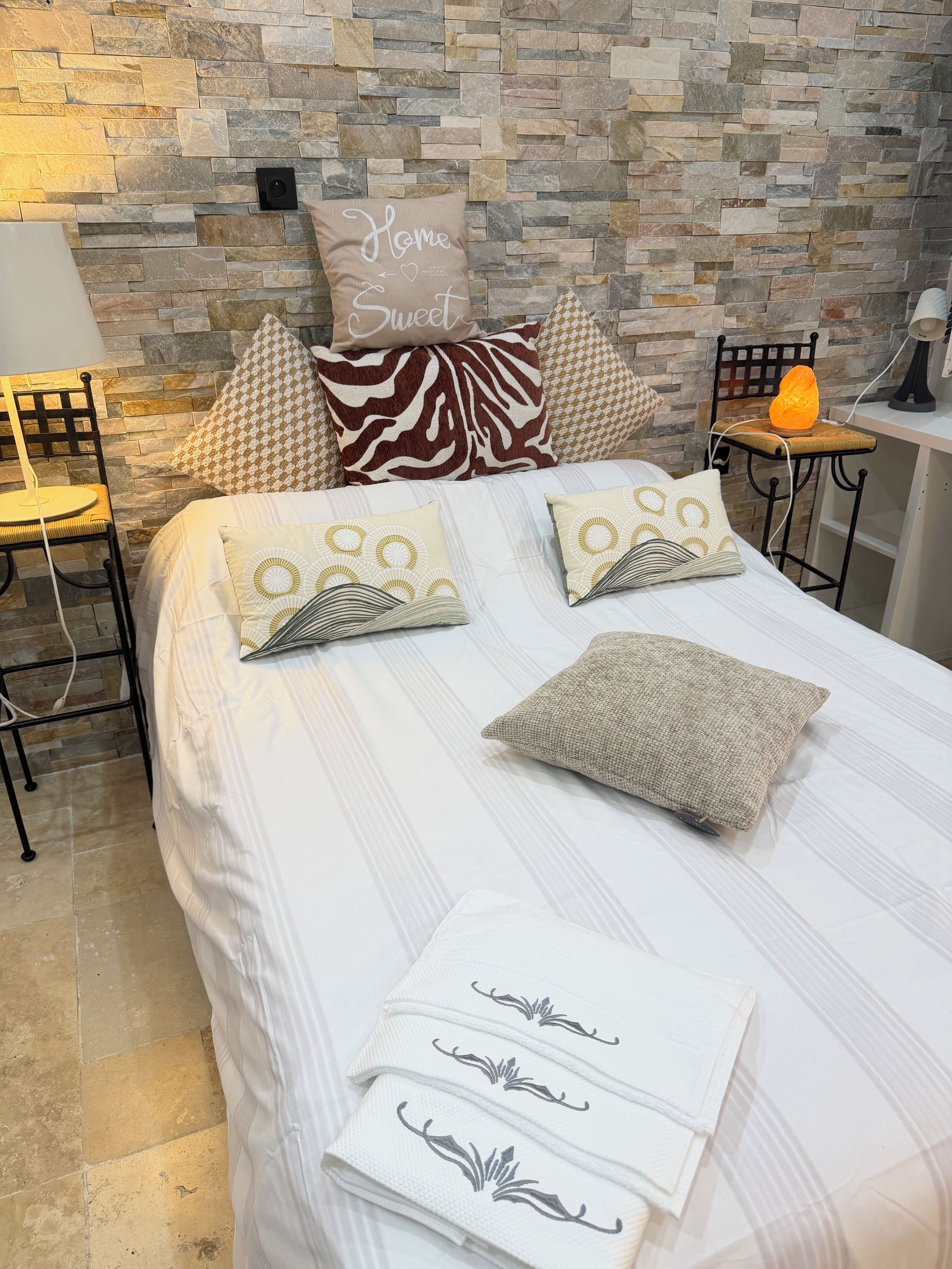 Una cama bien tendida con almohadones estampados, una pared con textura similar a la piedra y una silla con lámpara.