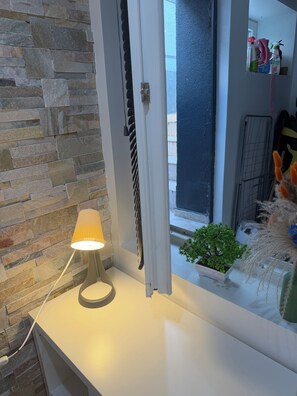Una habitación con pared de piedra, ventana con vistas, lámpara sobre una superficie blanca y una pequeña planta en el alféizar.