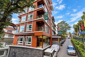 Un hotel de varios pisos con un restaurante en la planta baja, un estacionamiento y una calle con coches aparcados y vegetación.