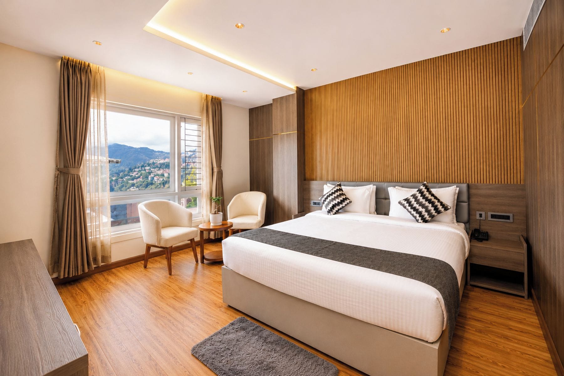 Habitación de hotel moderna con una cama grande, dos sillones, suelo de madera y vistas a un paisaje urbano.