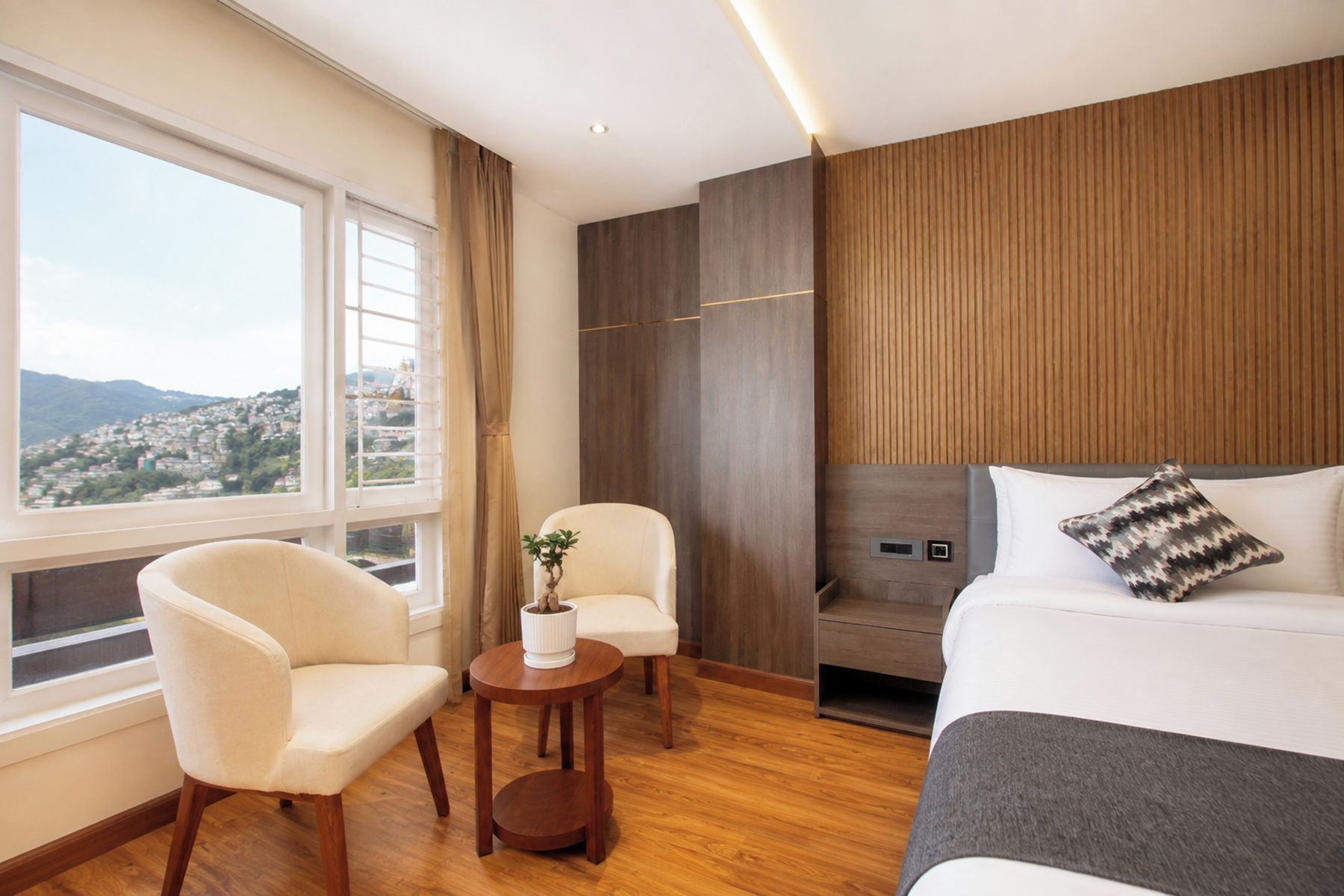 A hotel room with a bed, a chair, a small table with a plant, and a view of a cityscape through a window.