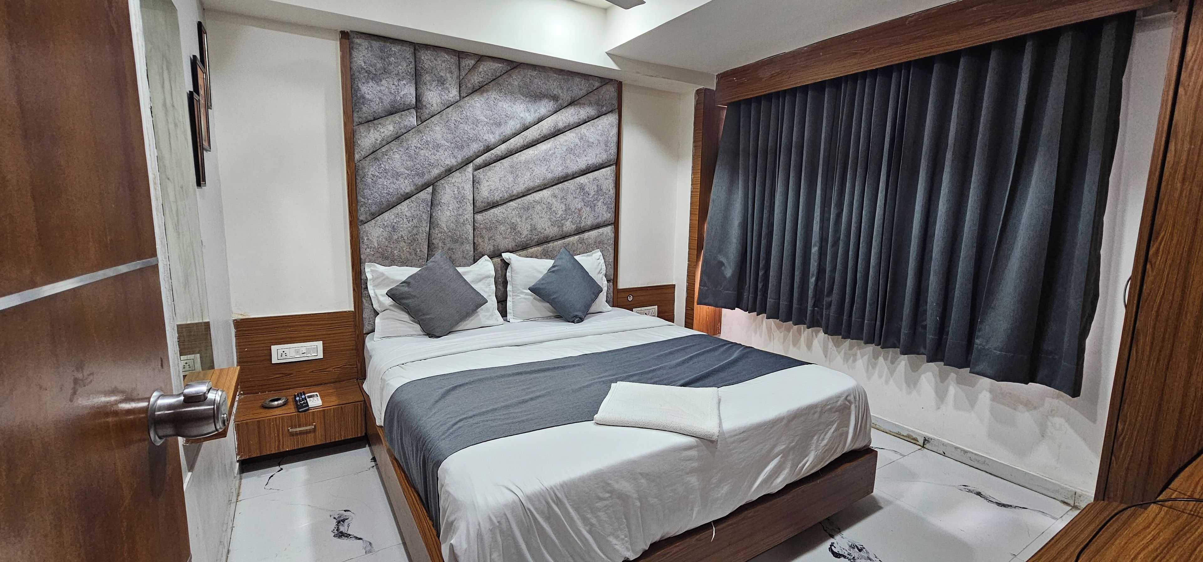 A bedroom with a bed, a nightstand, a wall-mounted headboard, and a window with curtains.