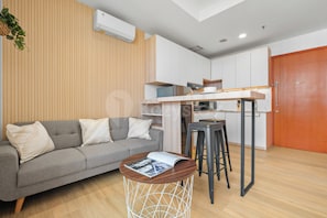 A compact living space with a kitchenette, a grey sofa with cushions, a small round table with a magazine, and a wooden floor. - Niren by Kozystay - Kuningan (South Jakarta)
