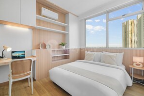 A modern bedroom with a bed, a desk, a chair, a window with a city view, and built-in shelves. - Niren by Kozystay - Kuningan (South Jakarta)