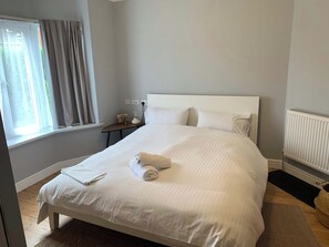 A neatly made bed with a rolled towel, a bedside table with a lamp, and a window with curtains. - One bedroom House close to Train station (Somerset)