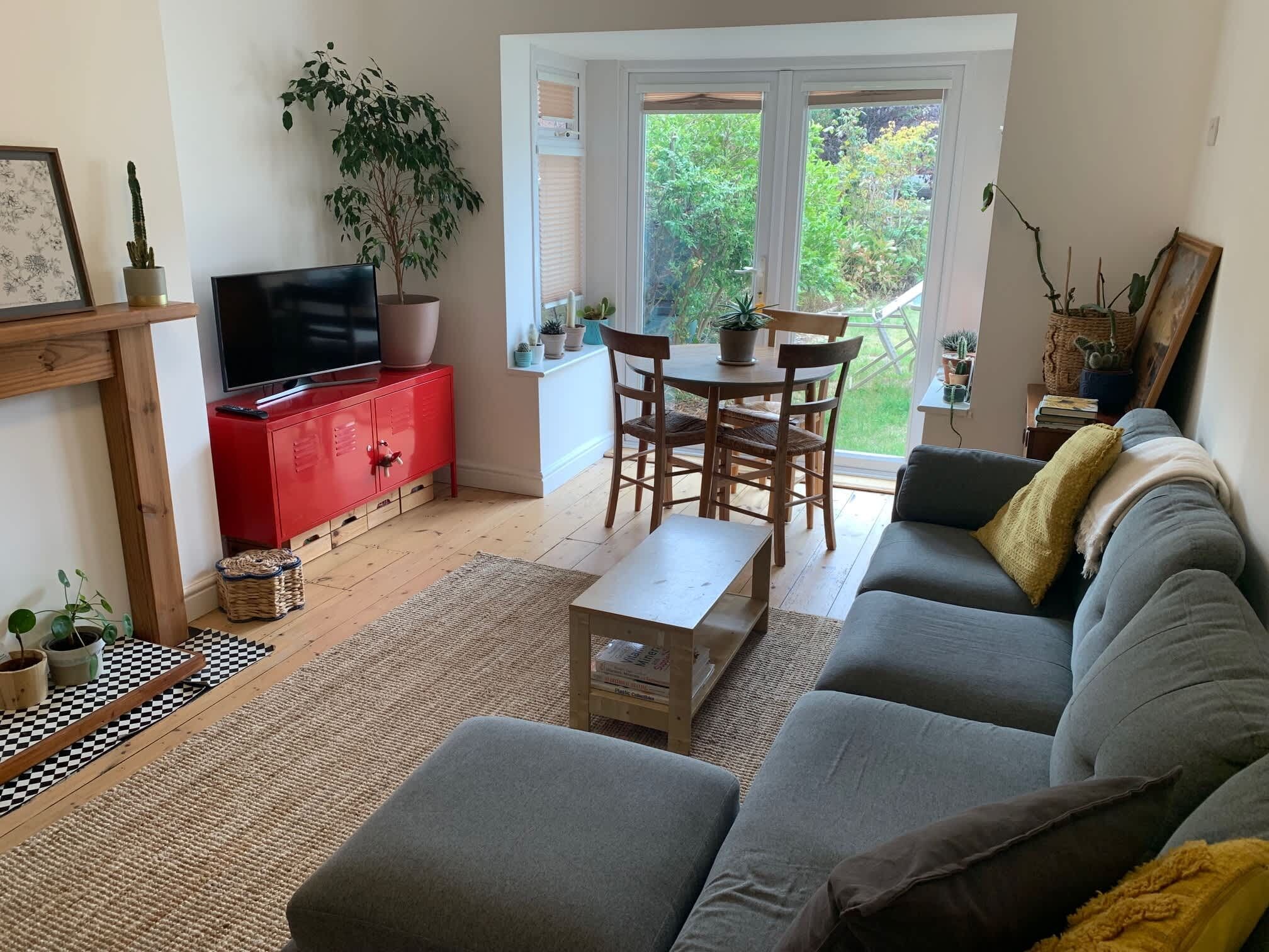 Una sala de estar con un sofá gris, un mueble de color rojo, un televisor, una mesa de comedor con sillas y vistas al verdor a través de una puerta de cristal.