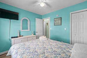 Quarto com parede verde-água, cama com colcha florida, cômoda com espelho e ventilador de teto.