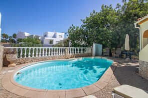 A swimming pool with a stone border, a white fence, and a view of buildings in the background.