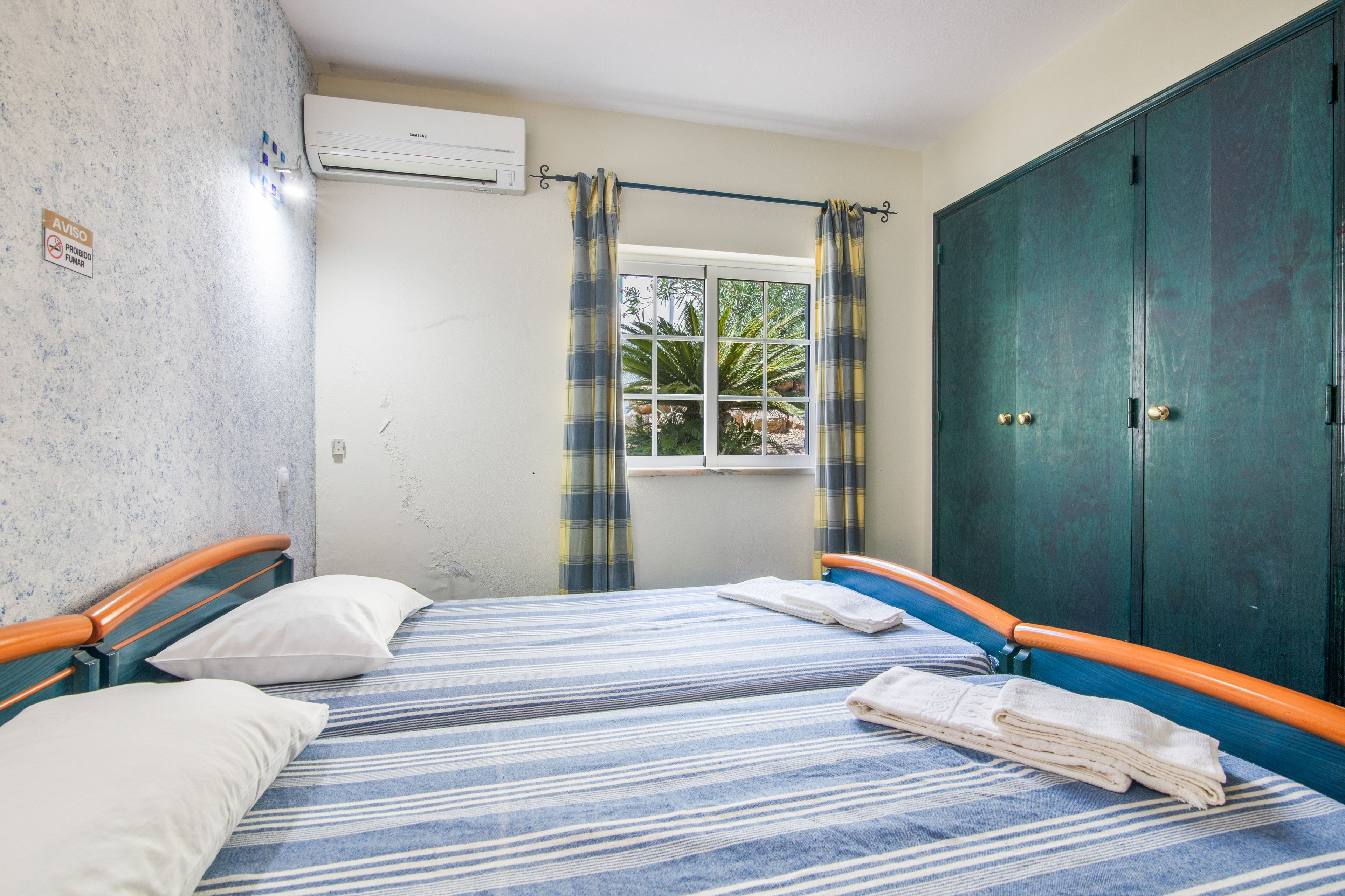 A room with two beds, a window with curtains, a wardrobe, and an air conditioning unit.