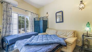 Un dormitorio con cama, sofá, ventana con cortinas, espejo en la pared y mesita de noche con lámpara.
