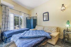 A bedroom with a bed, a sofa, a window with curtains, a wall-mounted mirror, and a bedside table with a lamp.