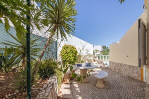 A courtyard with a stone table and chairs, surrounded by plants and a white building.