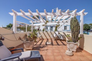A rooftop terrace with a pergola, potted plants, a seating area, and a view of a building with blue shutters.