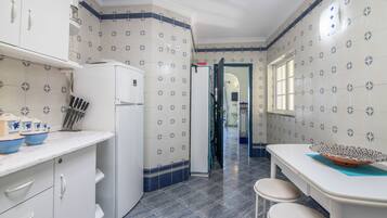 Una cocina con armarios blancos, un refrigerador blanco y un salpicadero de azulejos con diseño floral. Se observa una ventana en el lado derecho y una puerta que da a otra habitación.