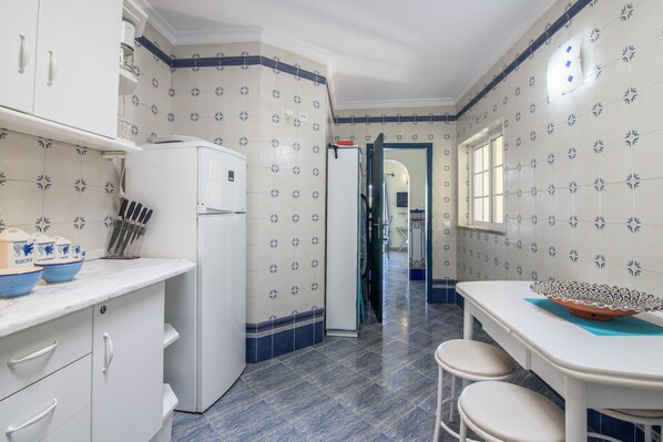 Una cocina con armarios blancos, un refrigerador blanco y un salpicadero de azulejos con diseño floral. Se observa una ventana en el lado derecho y una puerta que da a otra habitación.