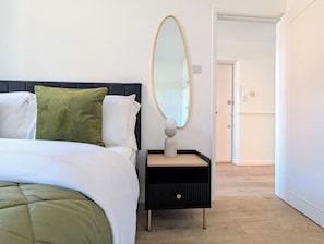 Siististi pedattu sänky, jossa on tumma pääty, yöpöytä peilillä ja ovi, joka johtaa toiseen huoneeseen.