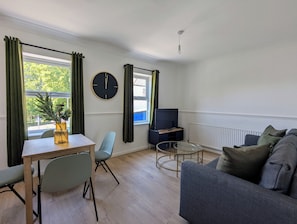 A modern living room with a sofa, a dining table with chairs, a TV stand, and a large window with curtains.