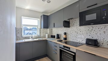 Ett modernt kök med grå skåp, en svart mikrovågsugn, en inbyggd ugn, ett fönster och en bänkskiva med nödvändiga köksredskap.
