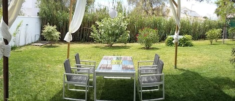 Een terras buiten met een tafel en stoelen, omgeven door een gazon en verschillende planten.