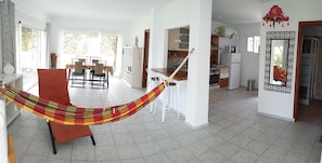 Ampla sala de estar com rede, mesa de jantar e cozinha ao fundo.