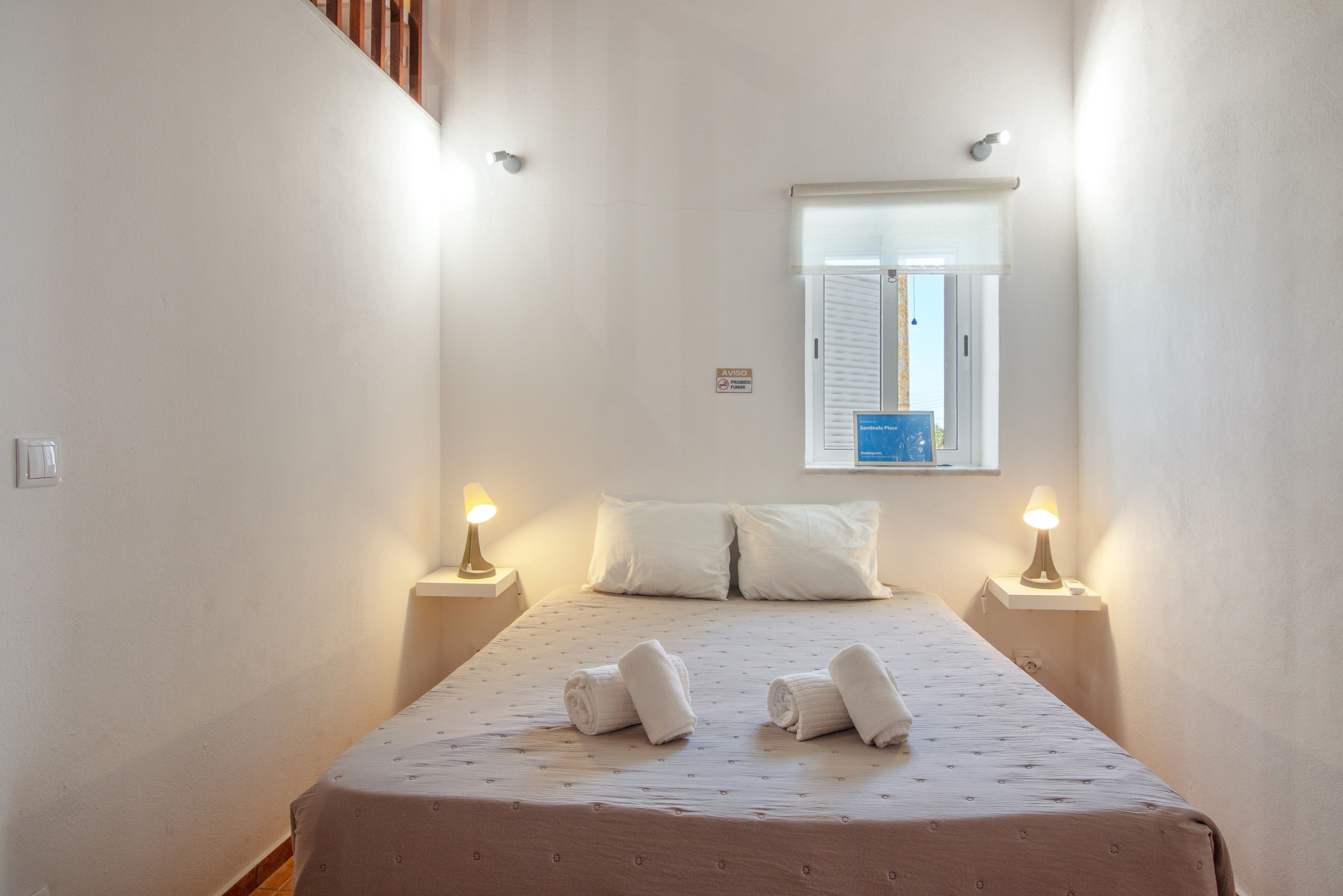 Habitación de hotel estrecha con cama, dos lámparas de noche, ventana con vistas y una mesita de noche pequeña.