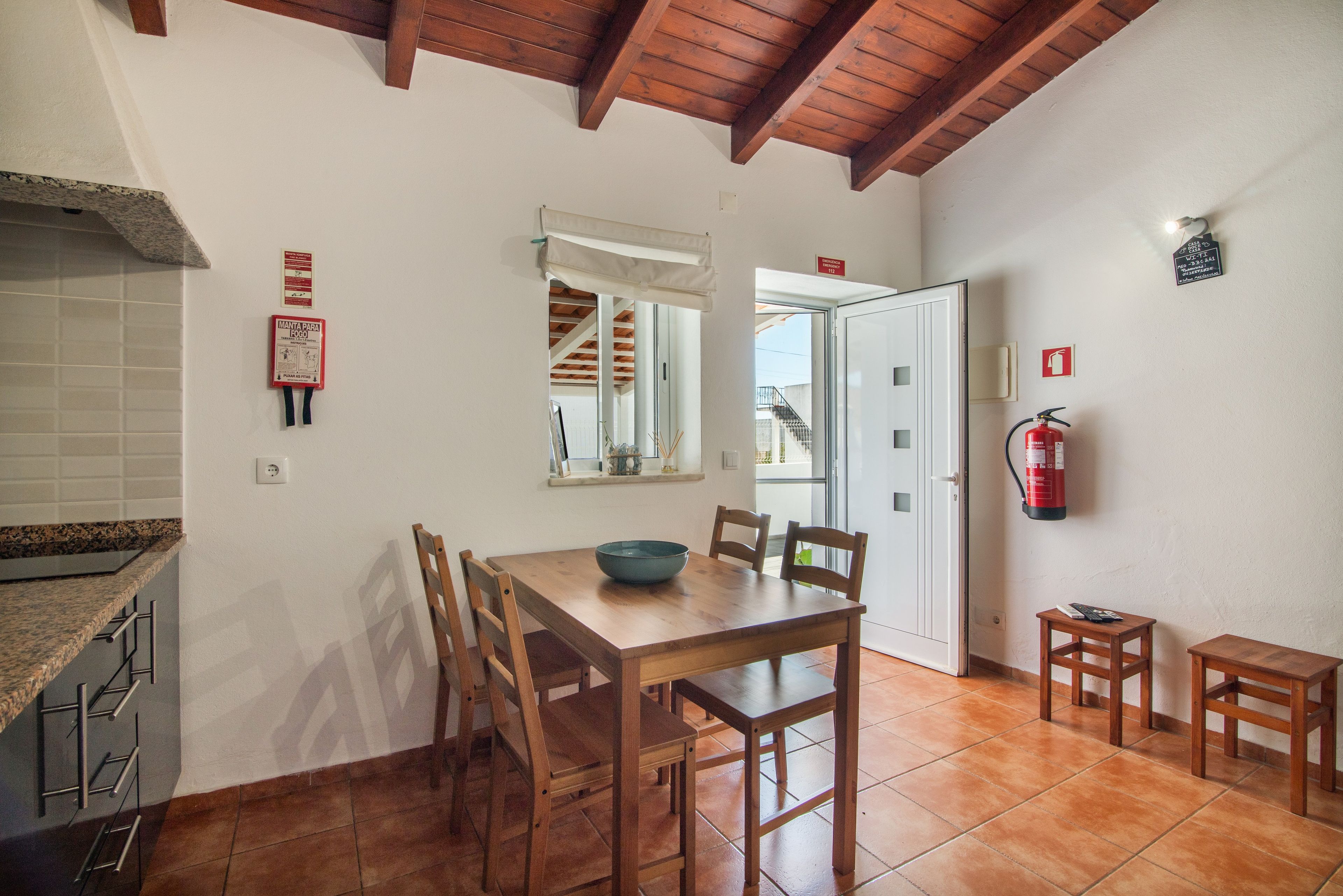 A dining area with a wooden table and chairs, a fire extinguisher mounted on the wall, and a window with a view of the outside.