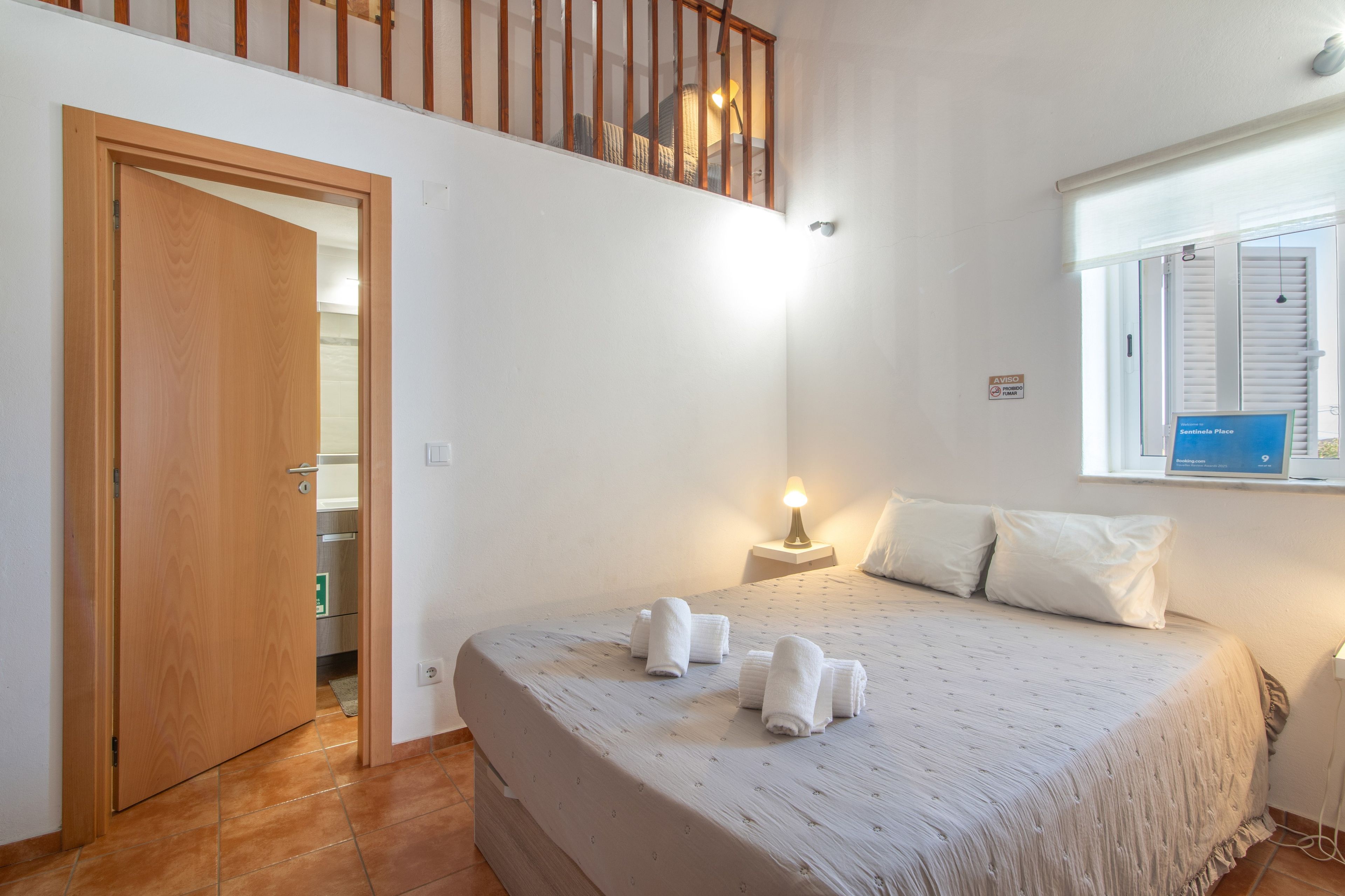 Een slaapkamer met een bed, een nachtkastje, een raam met jaloezieën, een deur en een trap.