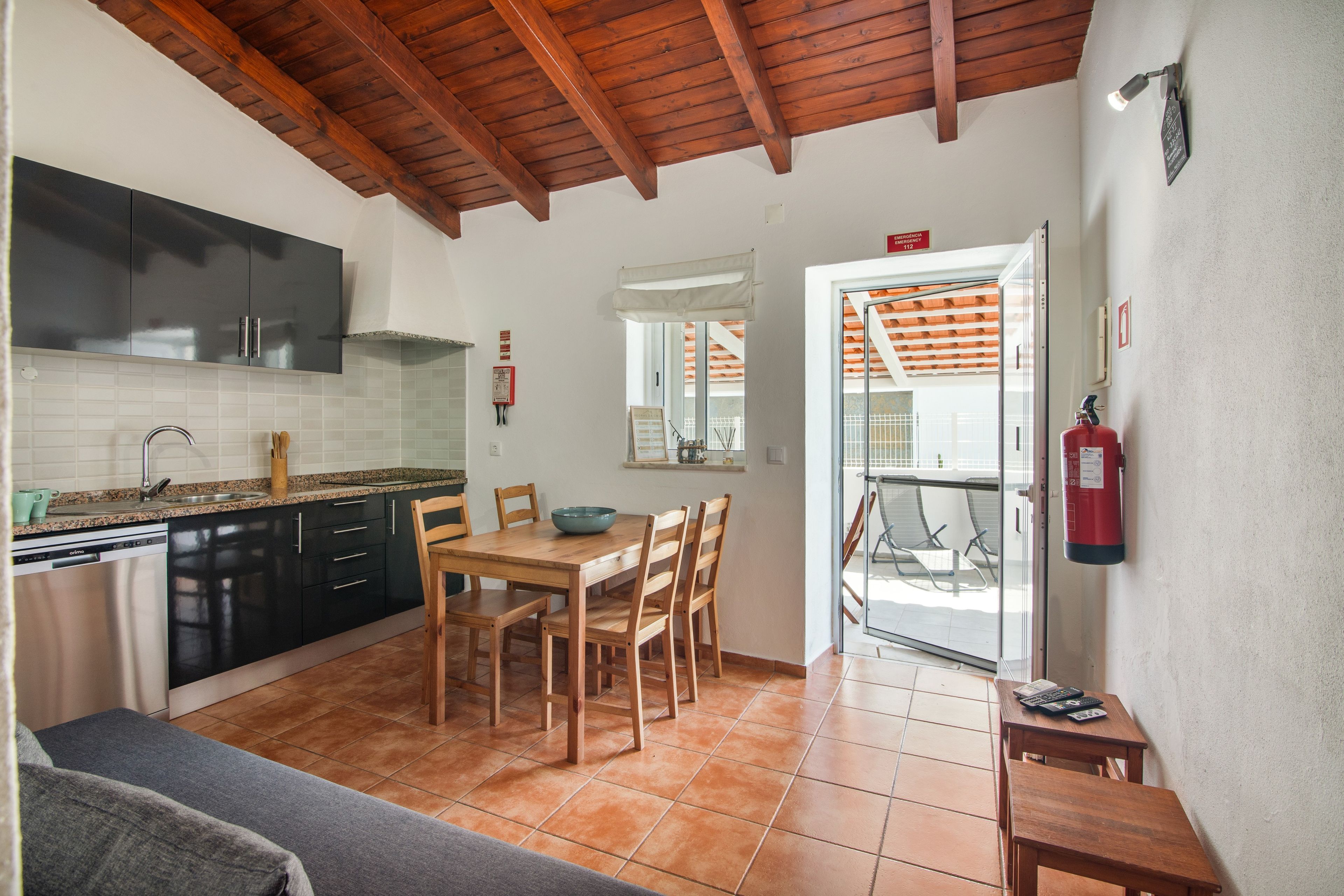 Cocina compacta con electrodomésticos modernos, zona de comedor con mobiliario de madera y señal de salida de emergencia visible.