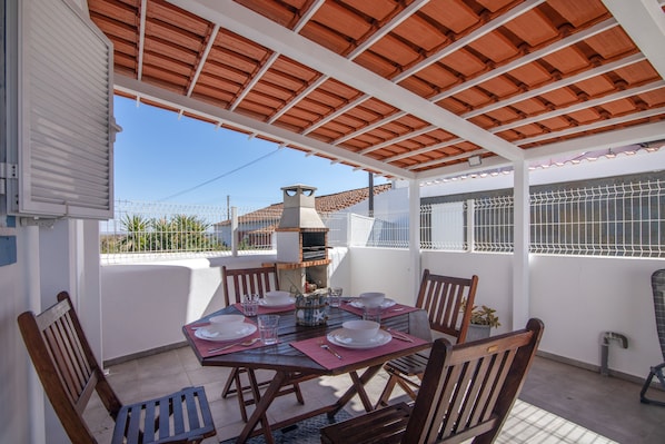 A covered patio area with a table set for dining, a barbecue grill, and a view of the surrounding area.