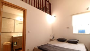 Une chambre à coucher avec un lit, une table de chevet, une fenêtre et une salle de bain visible à travers une porte ouverte.