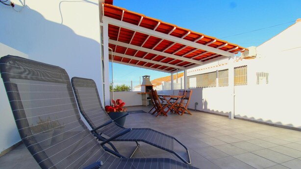 Un espace extérieur abrité, avec des chaises et une table en métal, un barbecue et un bâtiment blanc aux fenêtres grillagées.